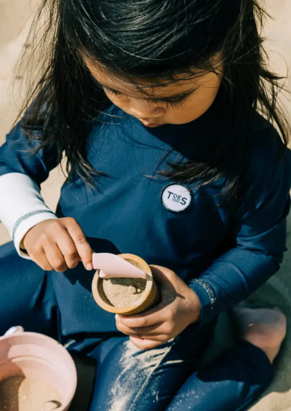 Child in a blue outfit playing with sand, scooping it into a small container. Sunlight casts shadows on the sand and the child's hair.