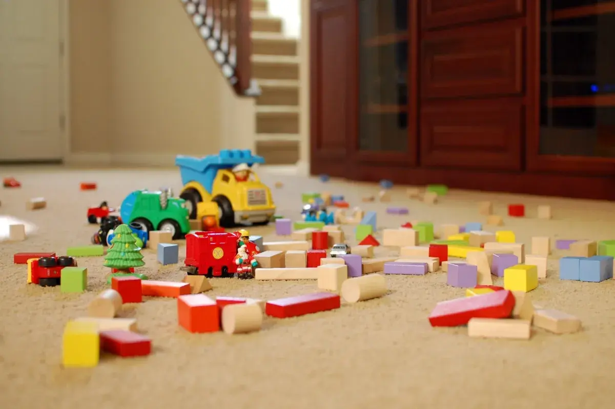 Toy cars and colorful building blocks scattered on a carpeted floor in a living room, with stairs and furniture in the background.
