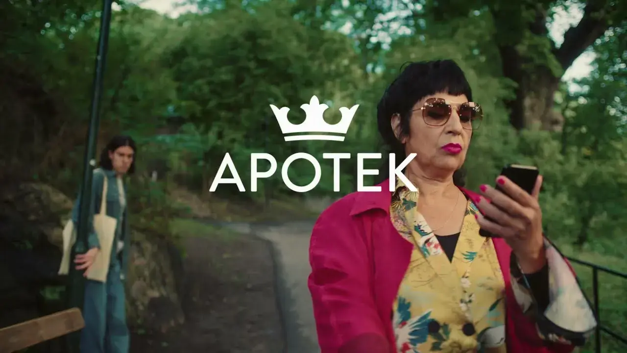 A stylish woman in sunglasses checks her phone while walking on a path in a park. A person with a bag follows in the background. "APOTEK" text overlay.