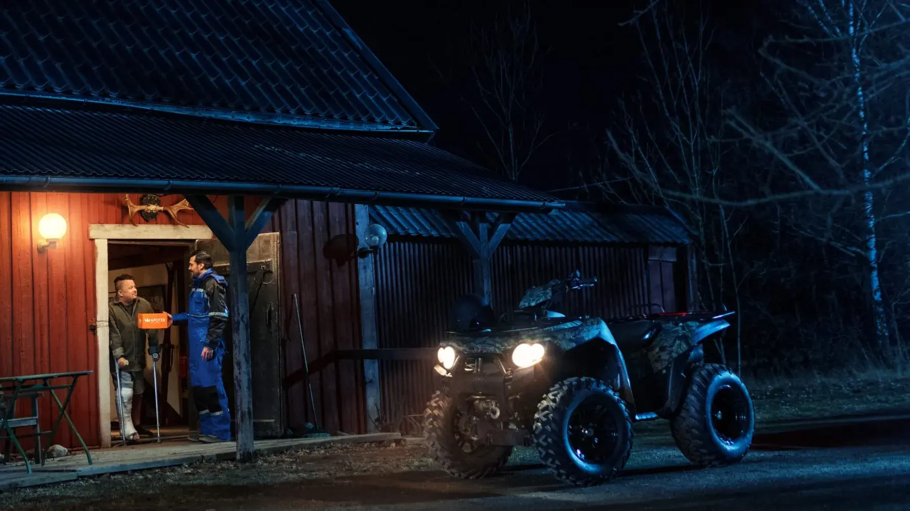 A person in blue overalls stands by an open barn door at night, talking to another person inside. An ATV with headlights on is parked nearby.
