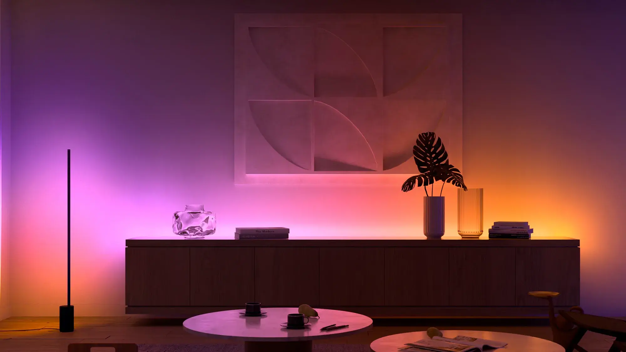 Modern living room with ambient purple and orange lighting, abstract wall art, and minimalist furniture, including a sideboard and round tables.