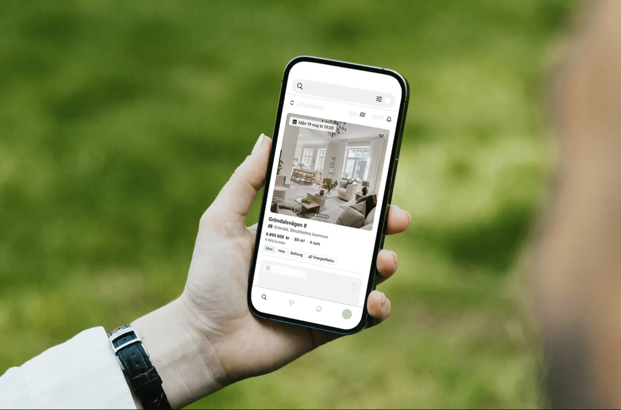 Hand holding a smartphone displaying a real estate app with a living room listing, set against a blurred green background.