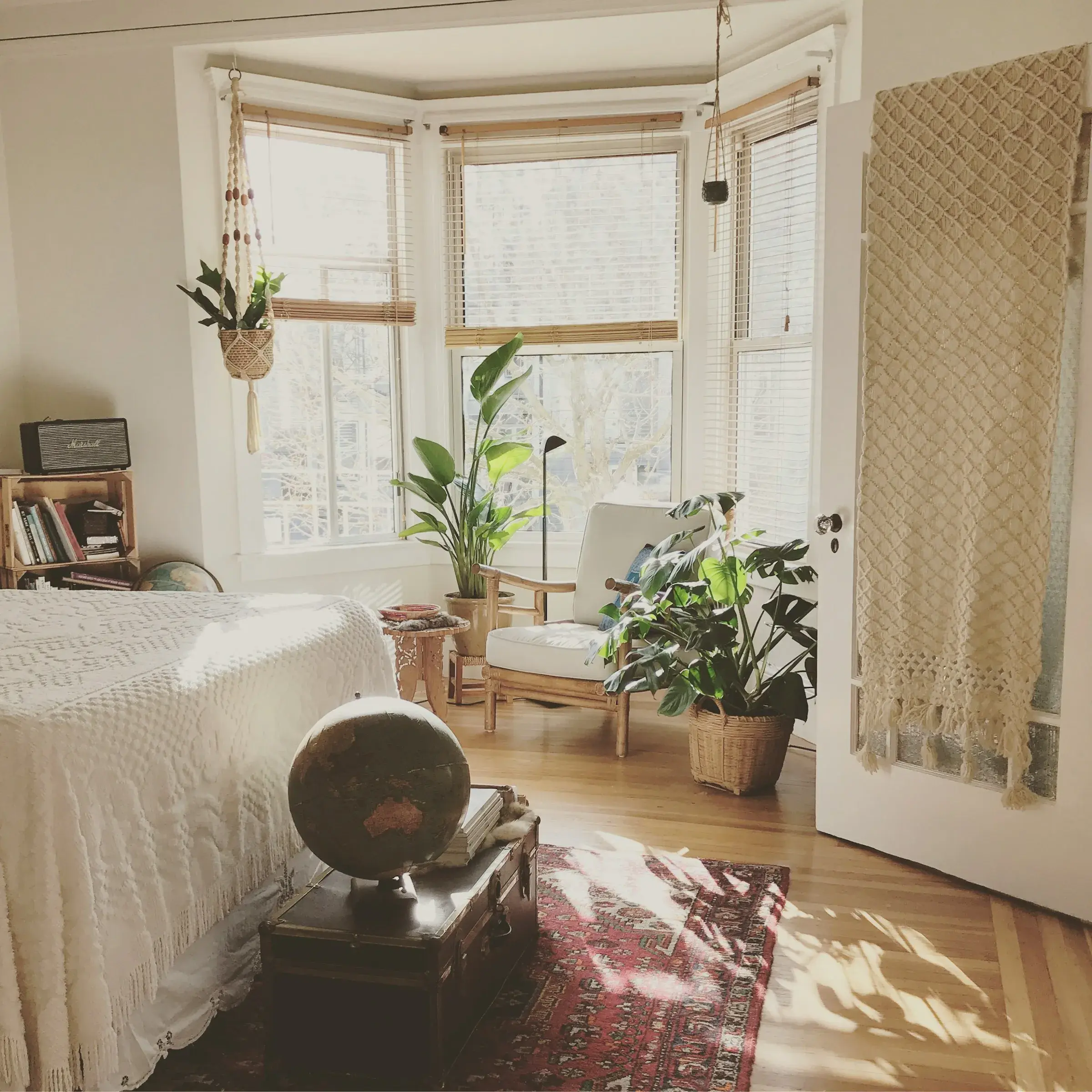 Sunlit bedroom with a cozy chair, plants, a bed with a white cover, a globe on a trunk, and a woven wall hanging on the door.