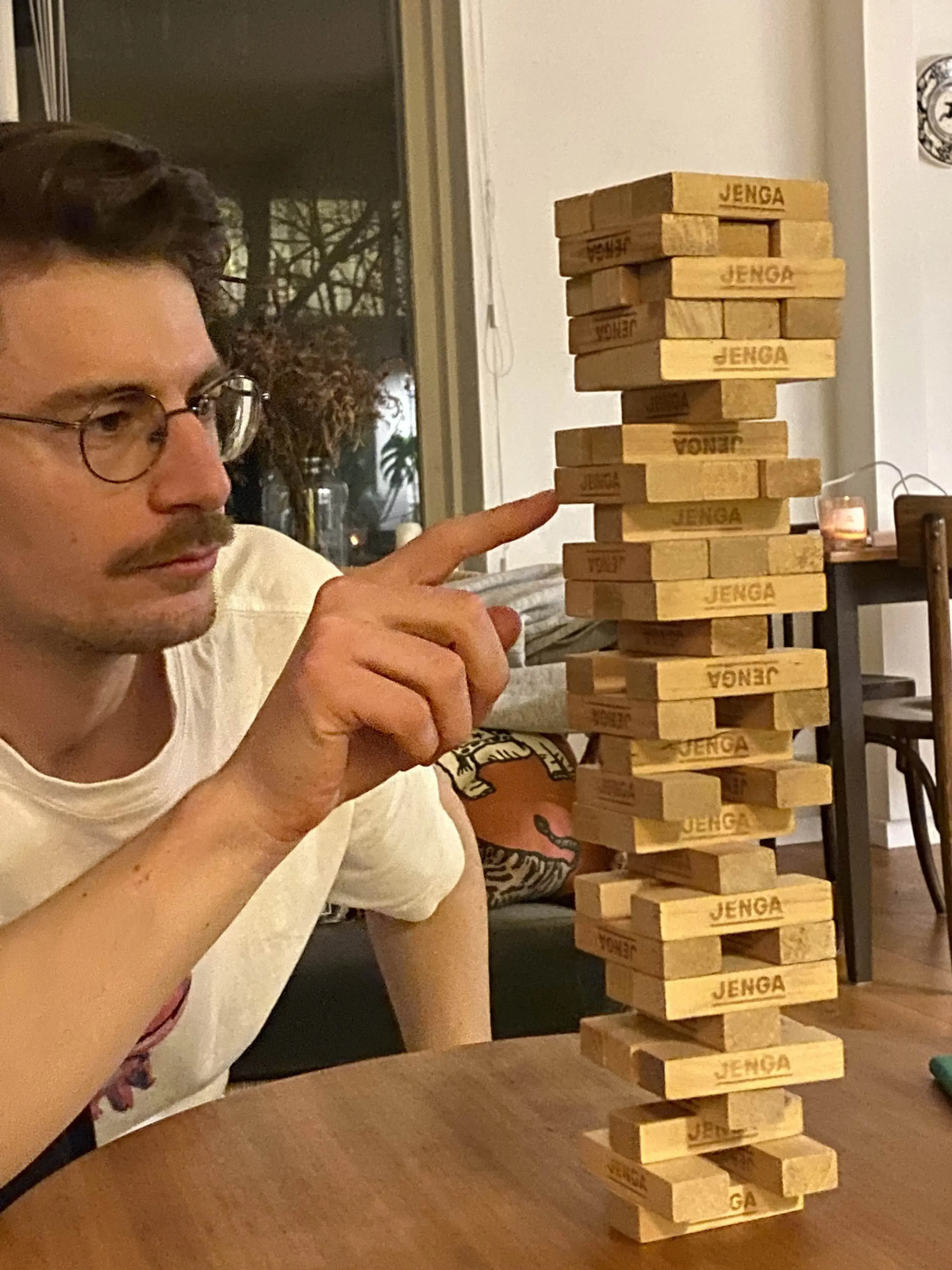 Person carefully removing a block from a tall Jenga tower on a wooden table, focused and wearing glasses.