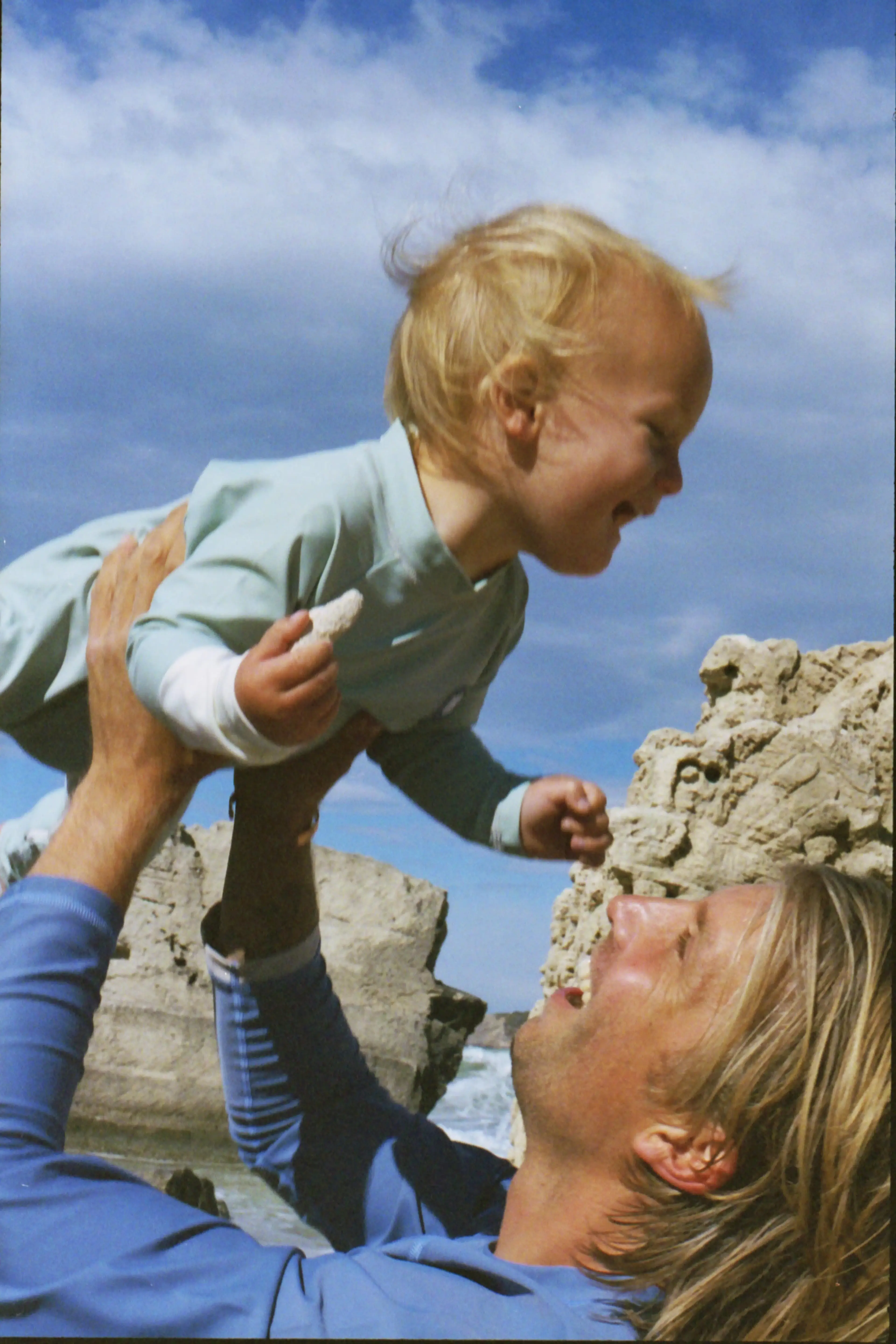 An adult playfully lifts a smiling toddler in the air by the beach. The sky is blue with scattered clouds.