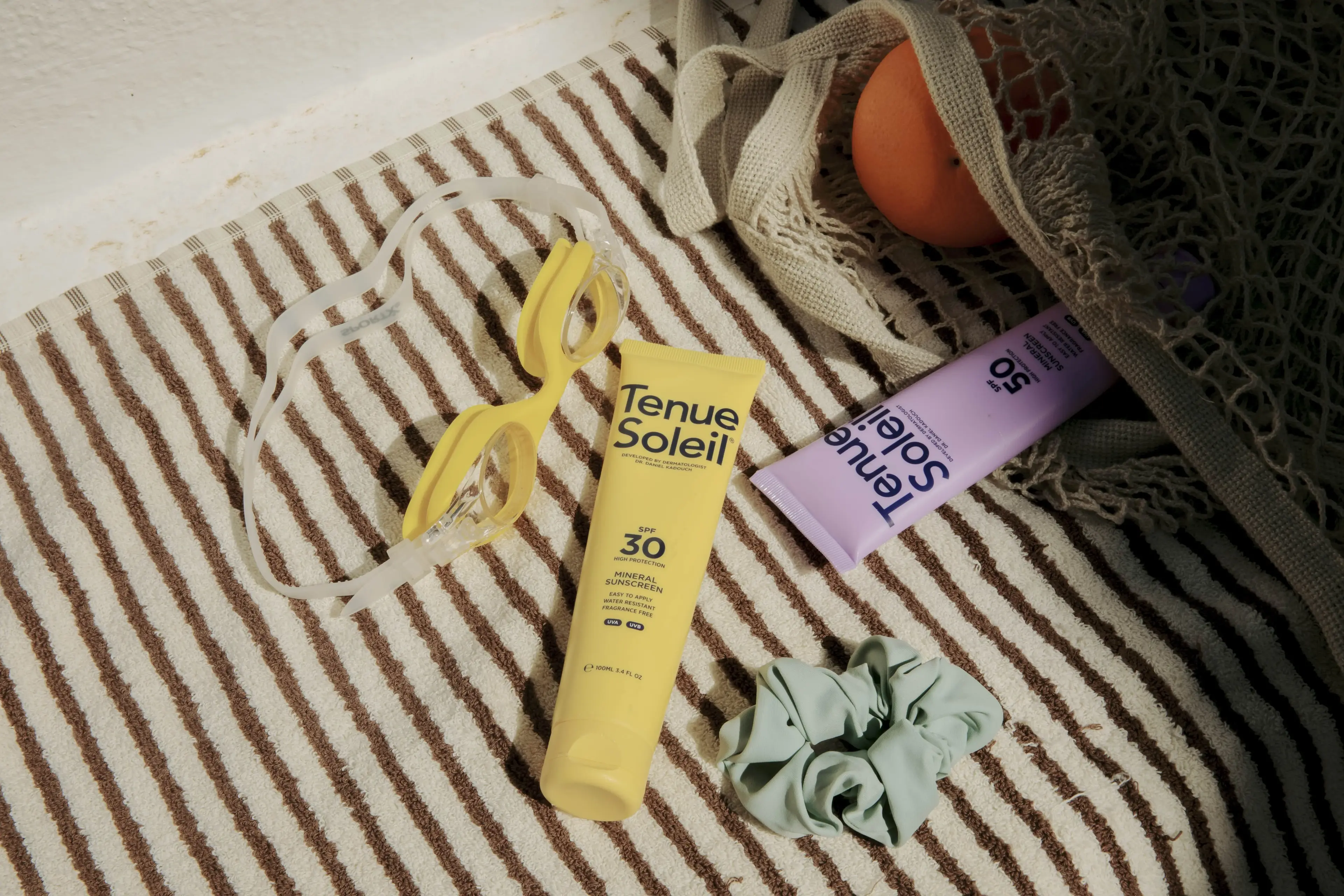 Sunscreen, goggles, scrunchie, and net bag on striped towel. Items suggest a beach or poolside setting.